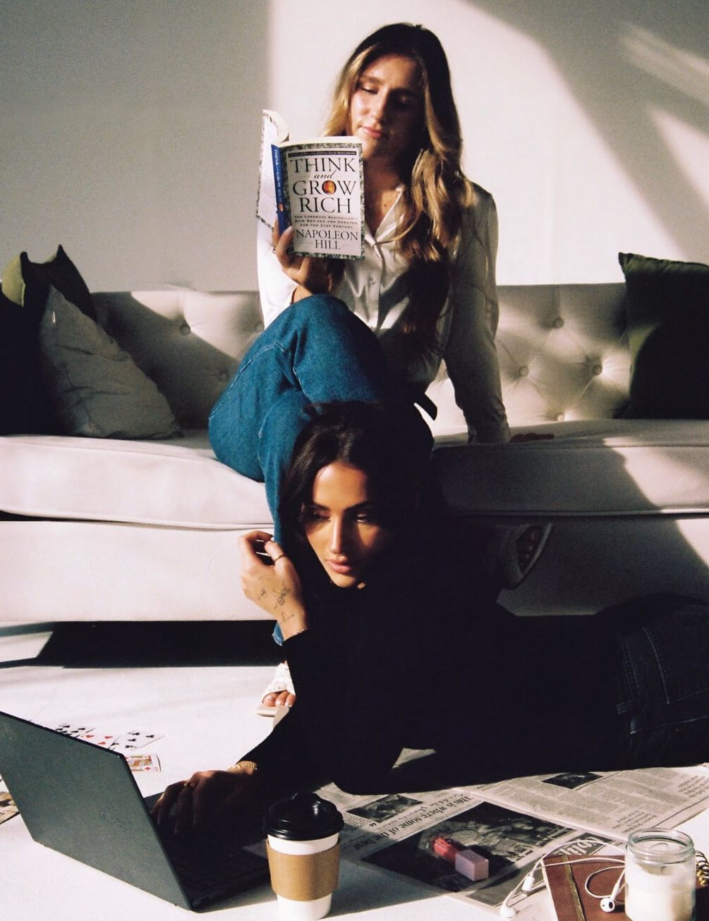 two women in a living room one is studying on the floor with open laptop while the other is sitting on a couch reading a book
